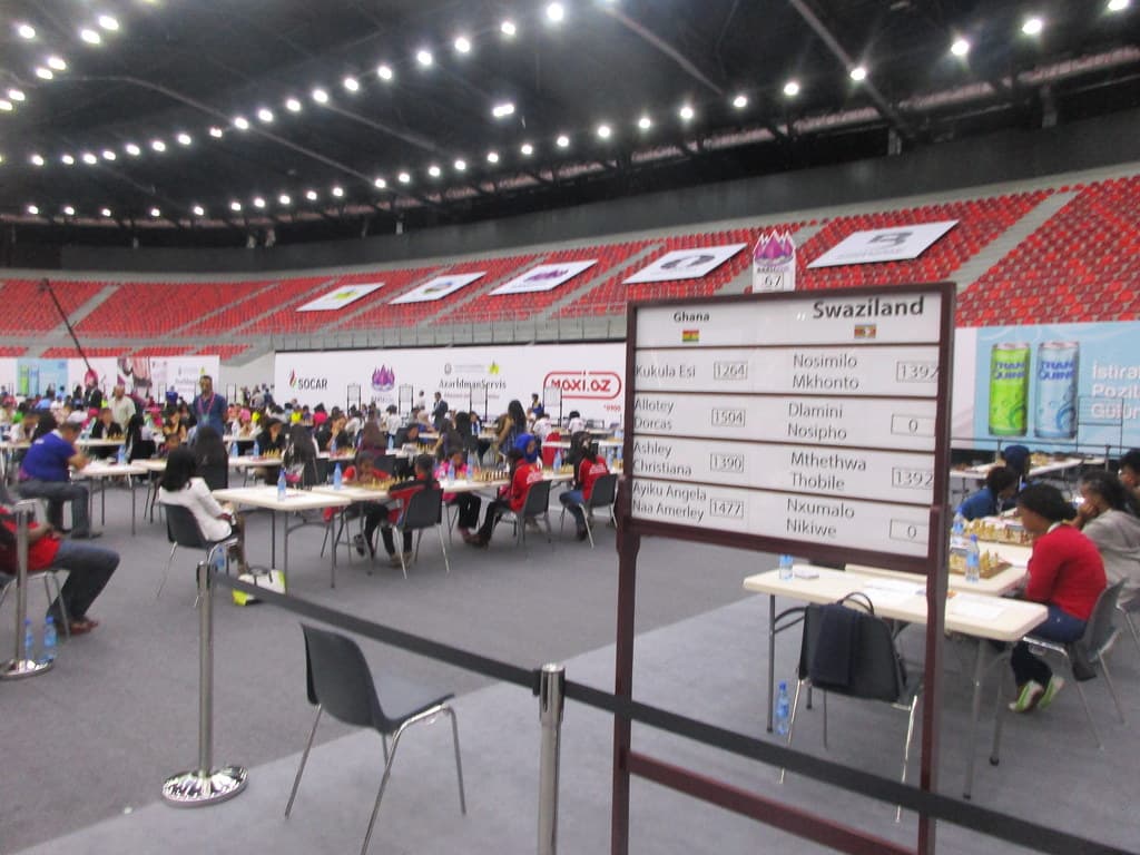 A Chess Olympiad hall, with a pairing board reading Ghana against Eswatini