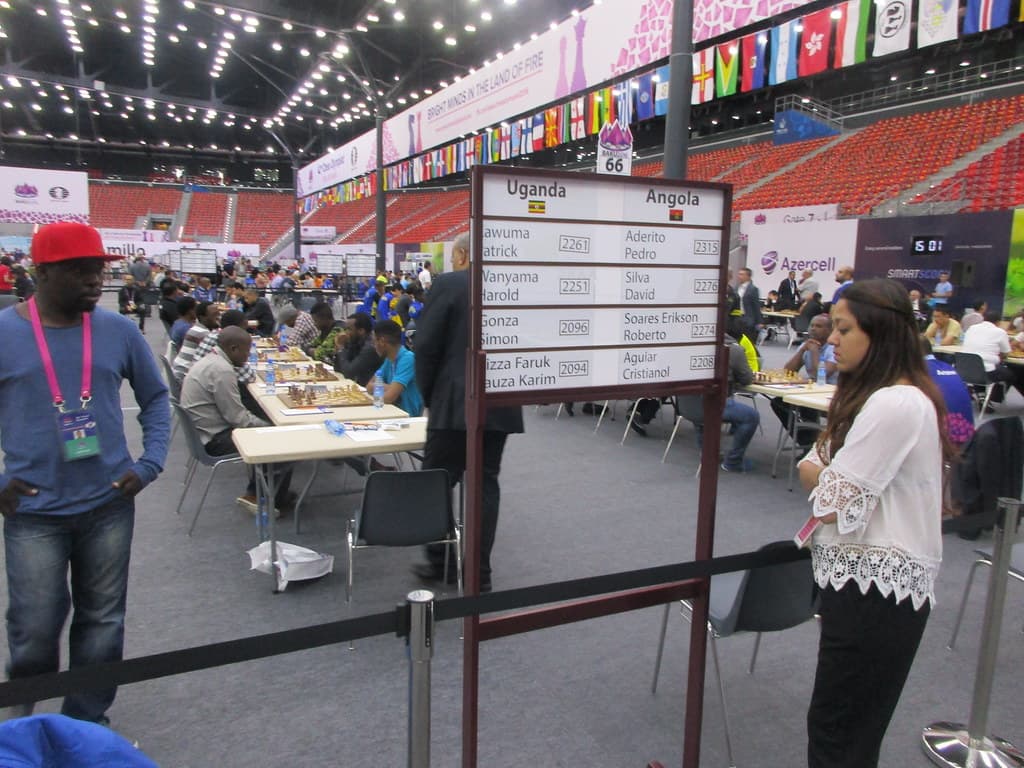 Players at the Chess Olympiad, with a pairing board reading Uganda against Angola