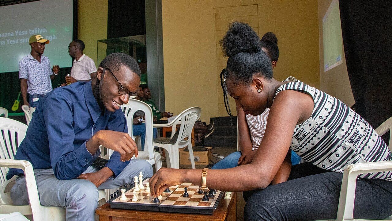 A man and a woman leaning over a board between two plastic chairs, both smiling
