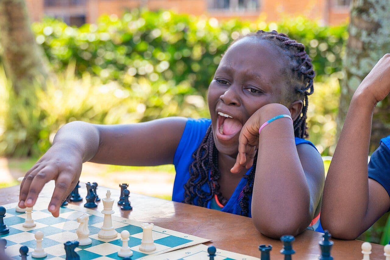 A young player reaching for a piece at an outdoor board, mid-laugh